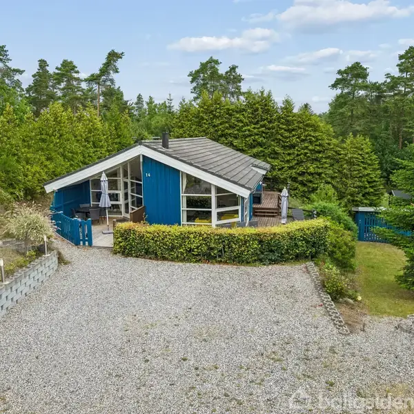 Blåt hus står stille omgivet af træer i en skovrig have. Foran huset er en grussti, og der er en terrasse til højre.