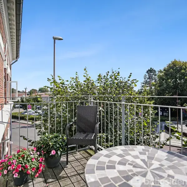 En lille, brolagt altan med en rund, mosaikmønstret bord og en sort stol. Omkranset af grønne planter og røde blomster med udsigt over vejen og træer i baggrunden.