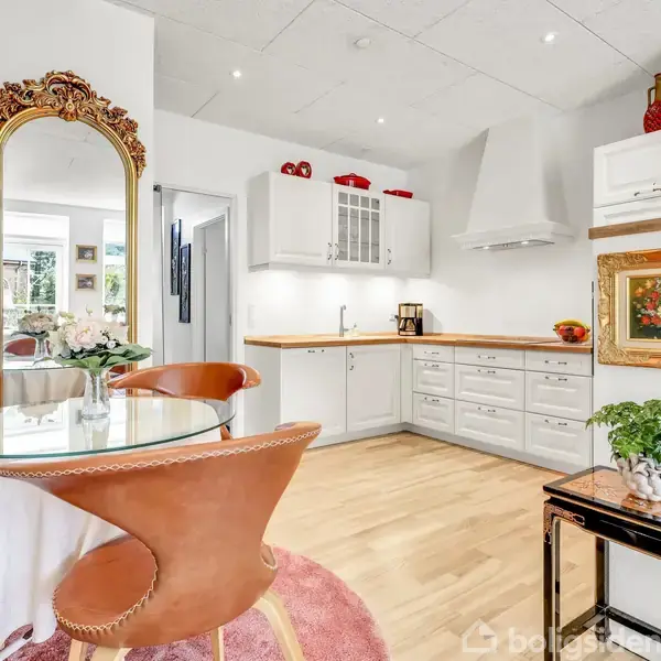 Et lyst køkken med hvide skabe og en glasplade spisebord med brune læderstole. Der er en stor dekoreret guldspejl ved siden af bordet og en plante på et lille bord.