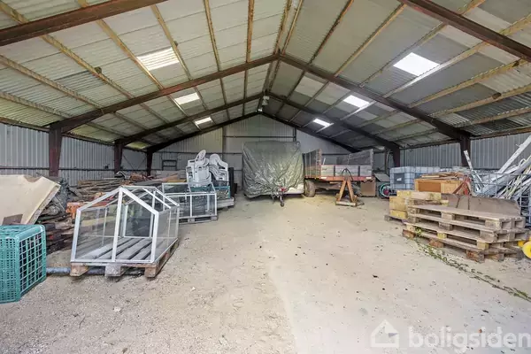 Et stort lagerhus indeholder stabler af paller, glasrammer og maskiner, delvist dækket af presenninger. Rummet er rummeligt med et højt, buet metaltag og betonvægge.