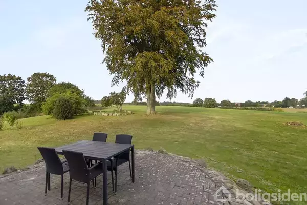 Et bord med fire stole står på en stenterrasse. Træet i baggrunden kaster skygger på den grønne græsplæne, omgivet af en landlig udsigt.