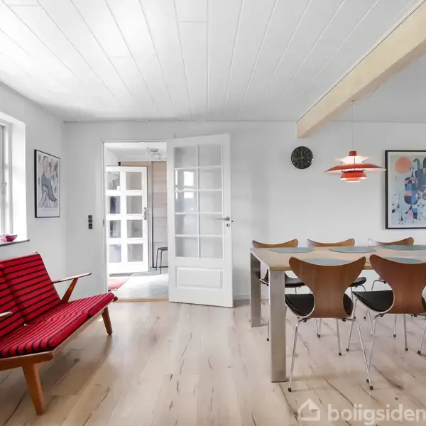 Sofa med rødt stribet stof står ved vinduet, mens spisebord omgivet af seks stole ses i lyse stue med hvidt loft. Væggene har kunstværker, og der hænger en rød lampe.