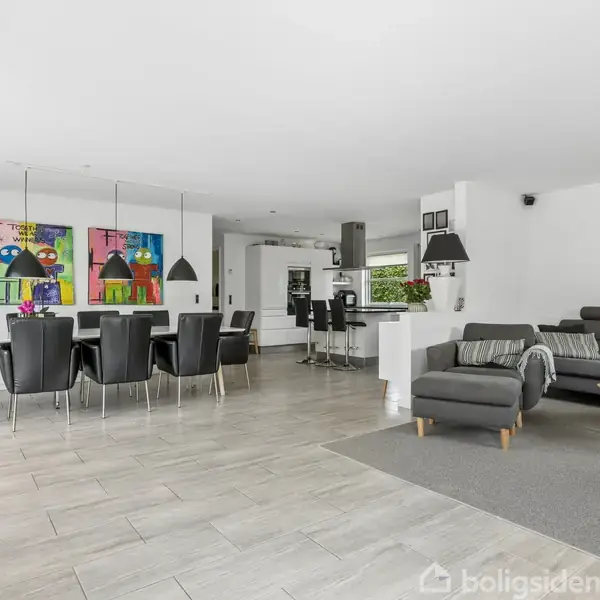 Spisebord med sorte stole står i en stue med lyse fliser, hvor moderne, koloristiske malerier hænger på væggen. I baggrunden ses en grå sofa og et åbent køkken.