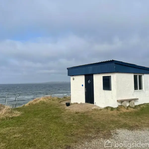 Et lille hvidt hus med fladt tag står på en græsplæne ved havet under en overskyet himmel. En bænk er placeret ved siden af bygningen.
