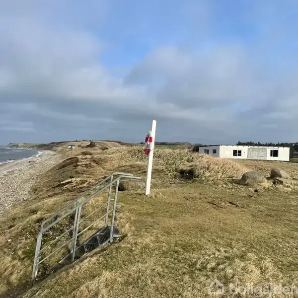 Græsbeklædt klippefremspring strækker sig langs stenet kyst, med bølger fra havet. Metaltrappe og mast med skilt står i midten. I baggrunden ses en hvid bygning og skove.