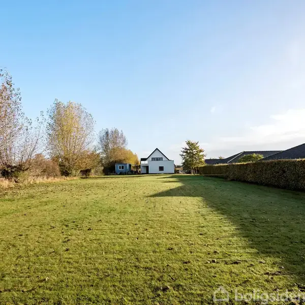 Hus står stille i baggrunden af en stor, grøn græsplæne med træer og buske rundt om, under en klar blå himmel.