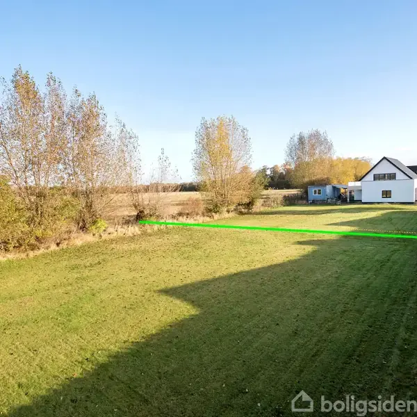Hus med hvid facade og sort tag står i haven. Græsplæne strækker sig foran, omgivet af træer og hække. Klar blå himmel. Ingen tekst fundet.