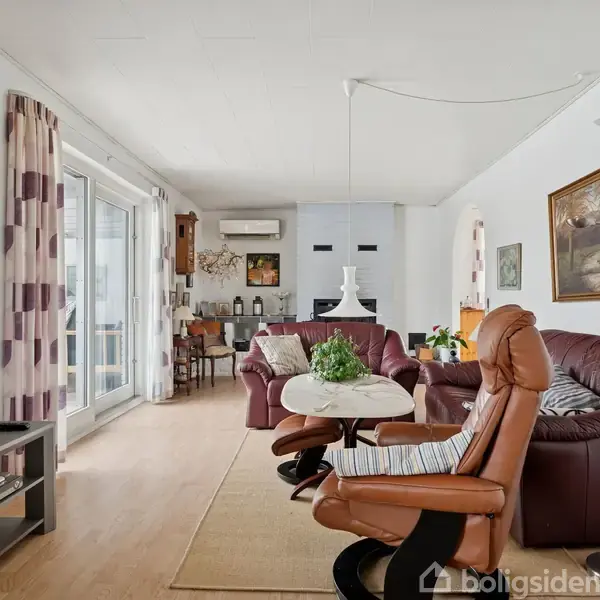 Stue med brun lænestol i forgrunden vender mod en sofa og et sofabord. Tv'et står til venstre, og store vinduer med gardiner slipper lys ind i rummet. En spiseplads anes i hjørnet.