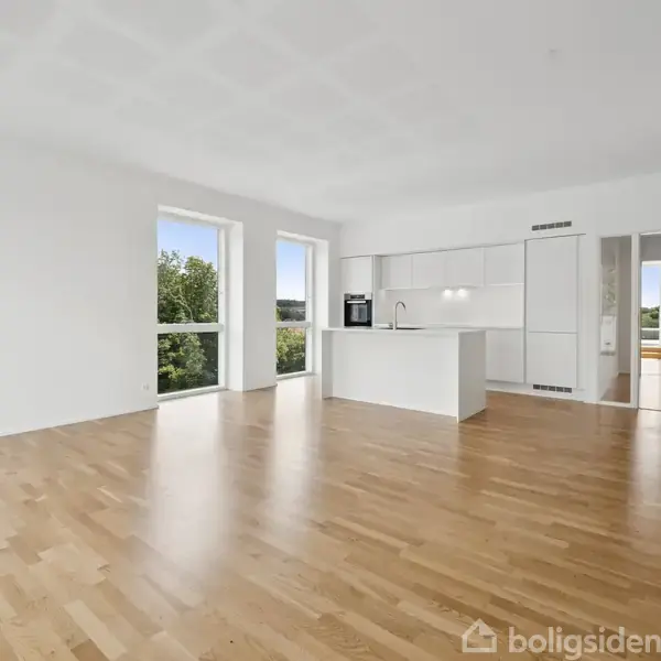 En lys gang med trægulv fører til et rum med store vinduer. Til venstre ses et åbent værelse, og for enden er en dør ud til en altan med udsigt til træer.