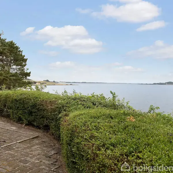 Et kystnært område viser en grøn hæk og flisebelagt sti langs vandet. Kraftige træer er til venstre, mens en rolig sø strækker sig i baggrunden under en blå himmel med spredte skyer.