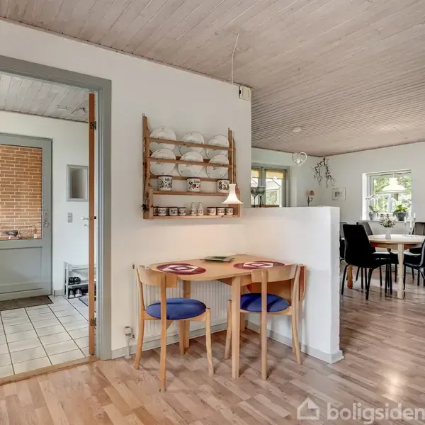 Et lille spisebord med to stole står ved en væg. Skåle og kopper er placeret på en hylde over bordet. I baggrunden ses et rummeligt køkken-alrum med spisebord.