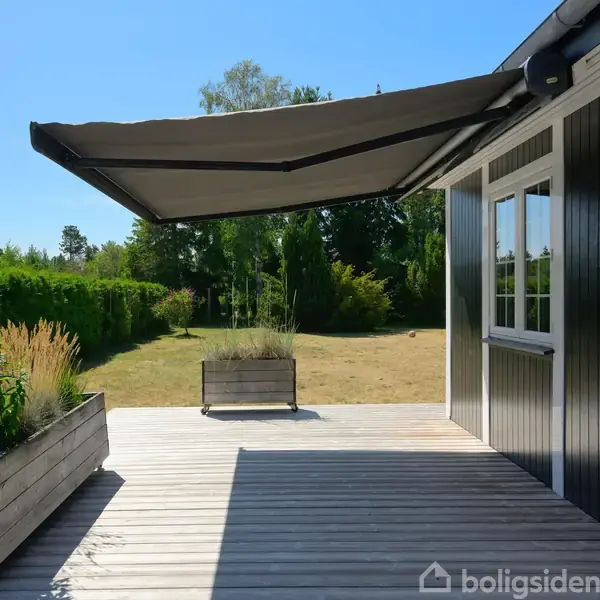 Markise udfoldet over en træterrasse ved et sortmalet hus. Planter i træbede pryder terrassen. Haven, fuld af grønne buske og træer, omkranser området. Klar blå himmel.