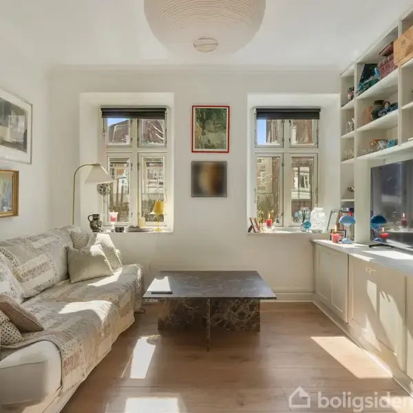Sofa står op af væggen med puder, omgivet af kunst på væggene. Bord foran sofaen. Til højre en reol med bøger og TV. Værelse er lyst med store vinduer.