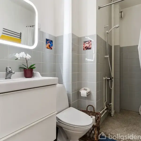 Badeværelse med hvidt toilet og vask under et oplyst spejl, omgivet af lysegrå fliser. En gul håndklæde hænger nær brusebadet. En orkidé står på vasken.
