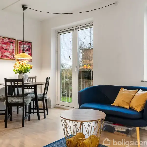 En stue med en blå sofa og gule puder ved siden af et rundt sofabord. I hjørnet står et spisebord med fire stole og en lampe hængende over det.