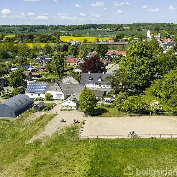 En ridebane med rytter i aktion omgivet af grønne marker og en landsby med huse og en kirke i baggrunden. Solpaneler er synlige på et tag.