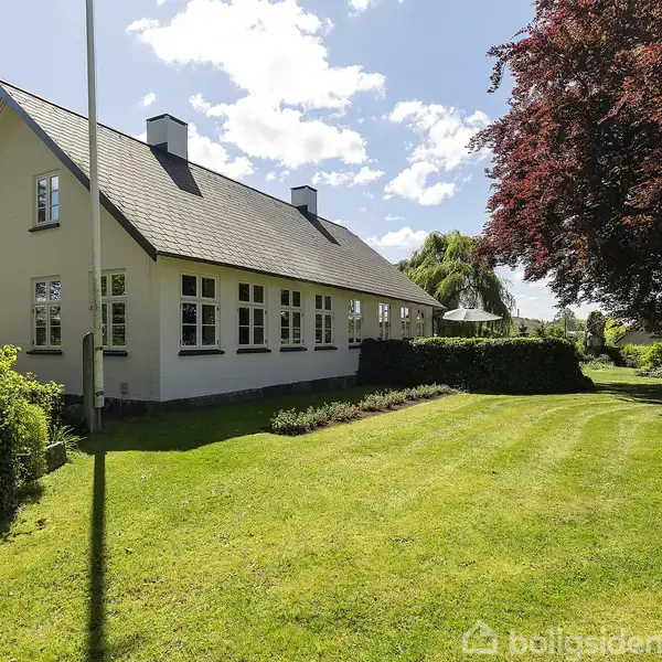 Hvidt hus med sorte tagsten står stille på en velplejet græsplæne. Omgivet af hække og et stort træ, under en solrig, skyet himmel.