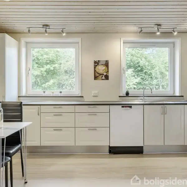 Køkken med hvide skabe og sort bordplade; bord med sorte stole til venstre; to vinduer med udsigt til grønne træer; loft med indbyggede spots. Flasker og kopper på bordet.