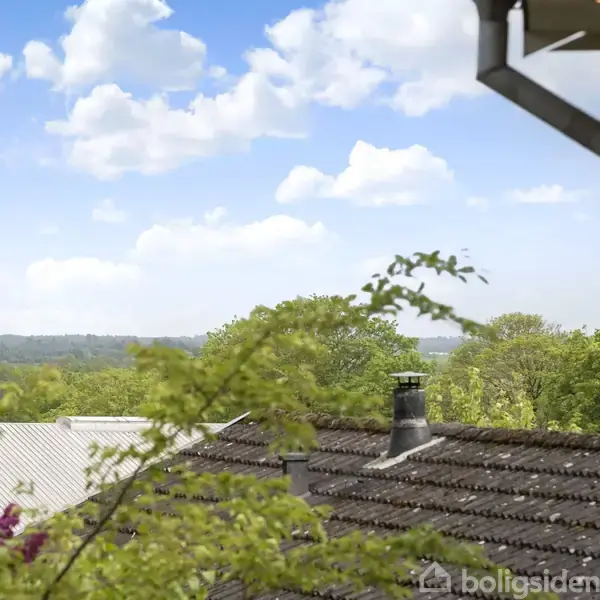 Tagrender hænger stille ved et hus, omgivet af frodig vegetation. I baggrunden ses tage af huse, med skorstene, og en udsigt over et grønt landskab mod en blå himmel med skyer.