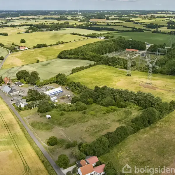 En luftfoto viser et landbrugsområde med marker og veje. En stor elledning tårner over landskabet ved siden af gårde og skovområder i en grøn, frodig landlig omgivelser.