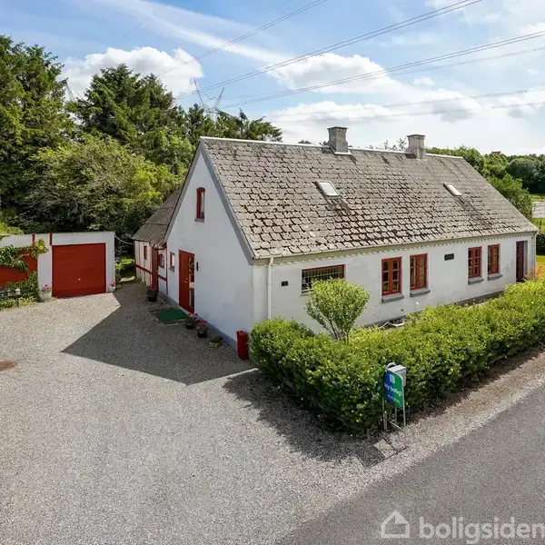 Et hvidt hus med røde vinduesrammer står på en grusvej. Foran huset er der en lille have med grønne buske. Bygningen har et skrånende tag.