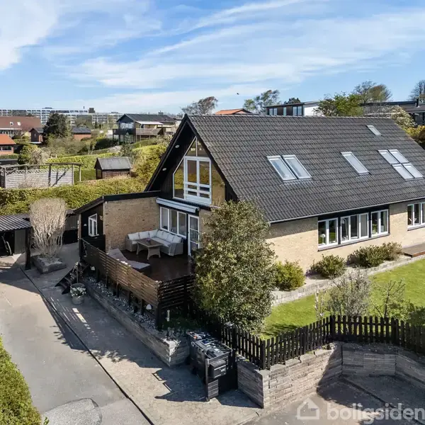 Et to-etagers hus med sort tag har store vinduer, en terrasse og en have. Beliggende i et boligområde med flere huse og grønne områder omkring.
