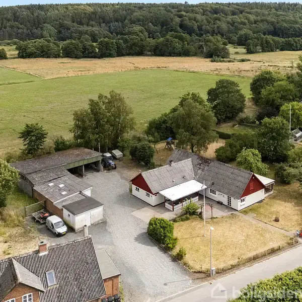 Hus omgivet af gårdsplads og mark; bilen er parkeret ved carporten, mens store træer skaber grænse mellem ejendom og landbrugsjord. Skovklædt område ses i baggrunden.