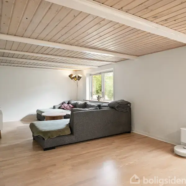 En grå hjørnesofa står i en lys stue med trægulv og træloft. Sofaen er mod en hvid væg med et vindue, og en ovn er tæt ved. En fladskærm hænger på væggen.