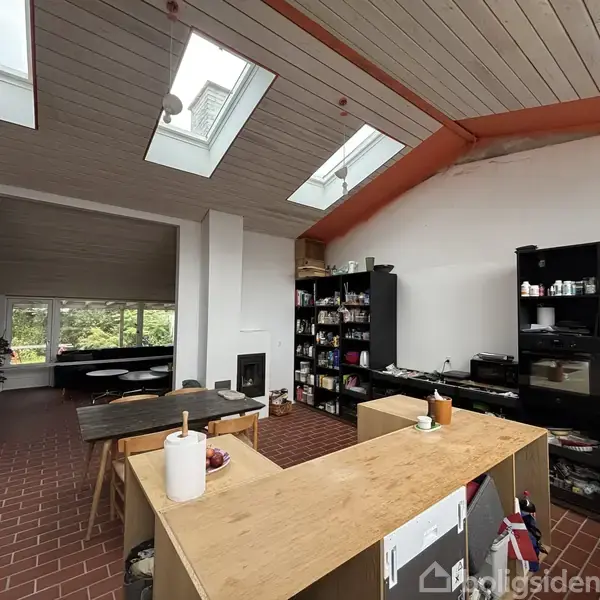 Et køkken med træbord og hylder, der indeholder forskellige køkkenartikler, står under et loft med ovenlys. I baggrunden ses en stue med grøn udsigt gennem vinduerne.