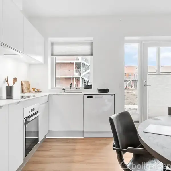 Hvidt køkken med indbyggede skabe og ovn, træspatler i en beholder, og skærebrætter står op ad væggen. Rundt spisebord med stole i forgrunden. Lys træbund og udgang til terrasse.