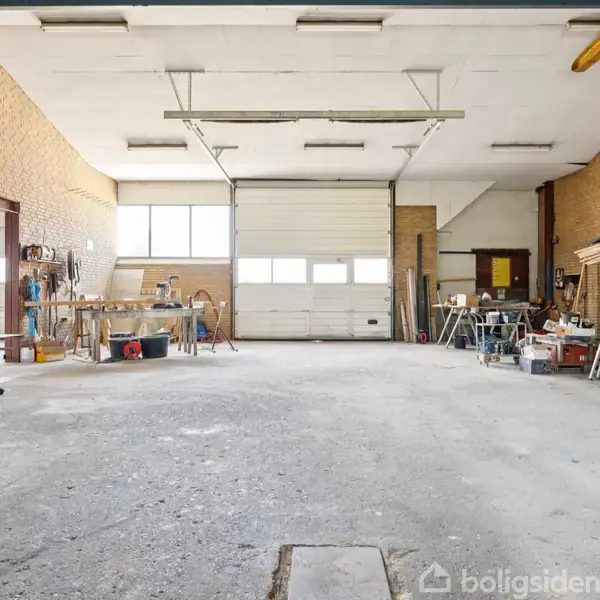 Et stort værksted med værktøj og materialer spredt omkring. Rummets vægge er af lyse mursten, og der er store garageporte samt arbejdsborde langs væggene.