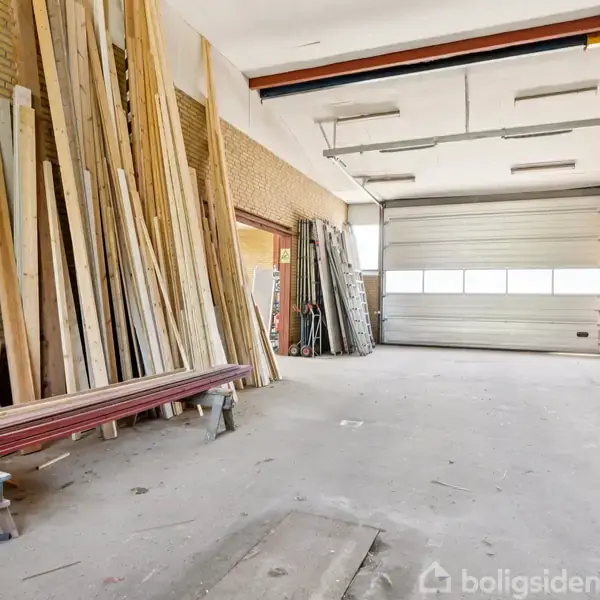 Træplanker står stablet langs murstensvægge i en garage med højt loft. Stort, åbent rum med en stor metalport i baggrunden og varierende byggeudstyr placeret rundt omkring.