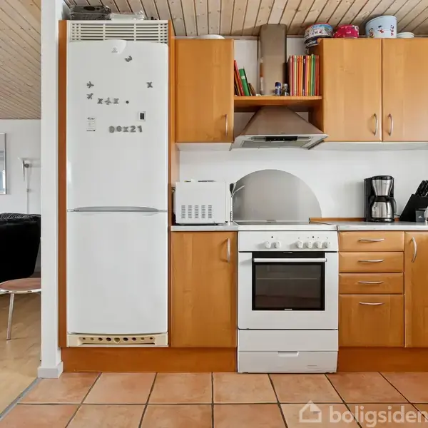 Et køkken med træskabe, hvor et komfur, en ovn og en mikroovn er placeret. Et hvidt køleskab står ved siden af. I baggrunden ses en sort sofa i stuen.