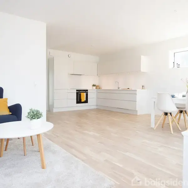 Stue med blå lænestol og gul pude ved hvidt sofabord. Køkkenet i baggrunden har hvide skabe. Rundt spisebord med fire hvide stole. Lyst trægulv og minimalistisk indretning.