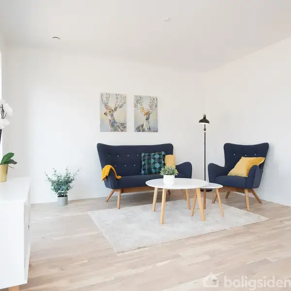 To blå sofaer med gule puder står i en lys stue med trægulv. Små borde og planter pynter rummet, mens vægkunst og en gulvlampe completerer det enkle, moderne look.
