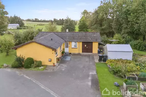 Et gult hus med mørkt tag står på en stor, grønt beplantet grund. En indkørsel fører til garagen. Omgivet af grønt landskab og flere træer i baggrunden.