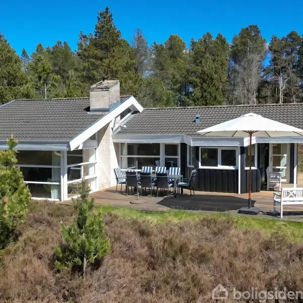 Et sommerhus med mørke paneler og en stor terrasse med havemøbler og parasol. Omgivet af skov, viser huset en afslappende naturlig atmosfære i en grøn, fredfyldt beliggenhed.
