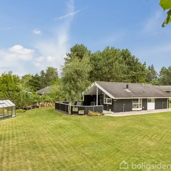 Sort træhus med terrasse står stille på grøn plæne omgivet af tæt skov. En lille glasdrivhus til højre beriger haven. Solrigt klarsyn fremmer fredfyldt stemning.