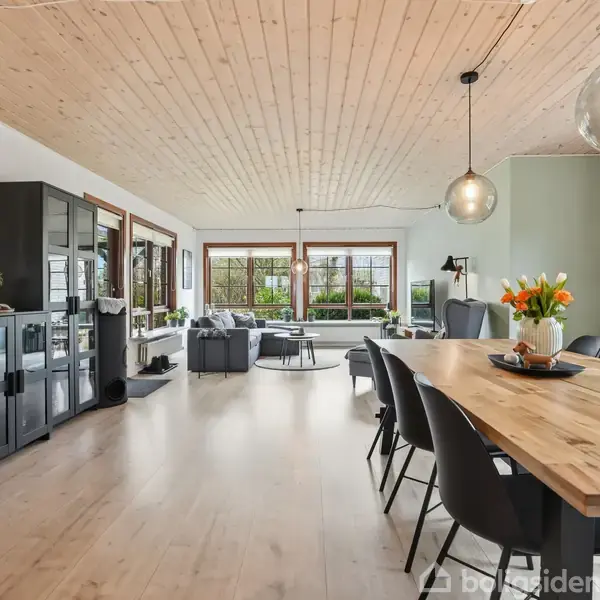 Stue med træbord omgivet af stole; store vinduer giver udsigt til haven. Moderne lamper hænger fra loftet, mens skænke og hylder pryder rummet. Blomster pynter bordet.