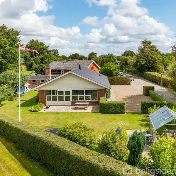 Et hvidt hus står i midten omgivet af grønne hække og træer, med en flagstang til venstre. Et drivhus ses foran, med en blå himmel og skyer ovenfor.