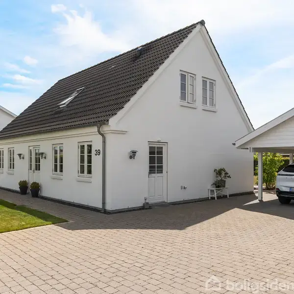 Hvidt parcelhus med sort tegltag står på en flisebelagt indkørsel. Til højre er en carport med en parkeret bil. Husnummeret "39" er synligt på facaden.