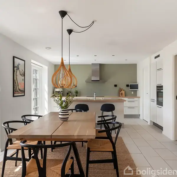 Spisebord med sorte stole står i et lyst køkken. Over bordet hænger to trælamper. En åben dør fører ud til en terrasse med parasol. Billede på væggen.
