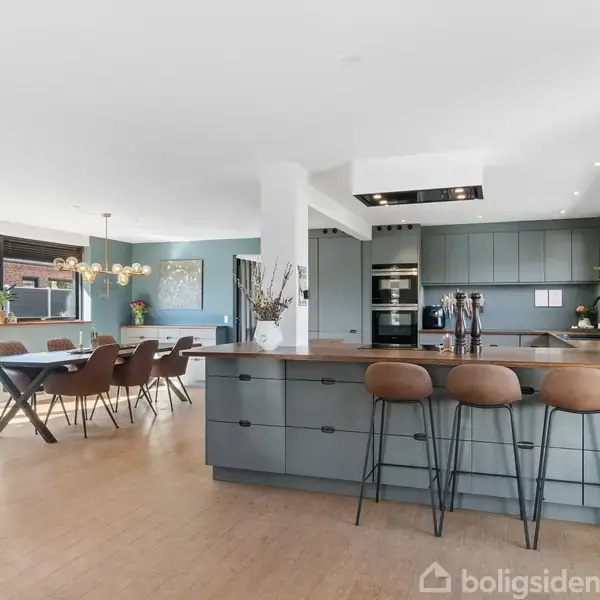 Et moderne køkken og spiseområde med træmøbler og grønne skabe. Køkkenøen har barstole, og i baggrunden ses et spisebord med stole. Store vinduer giver naturligt lys.