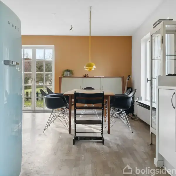 Et moderne køkken med et lyseblåt SMEG-køleskab til venstre, et langbord med sorte stole i midten, og en hvid skænk foran en orange væg. En gul lampe hænger over bordet.