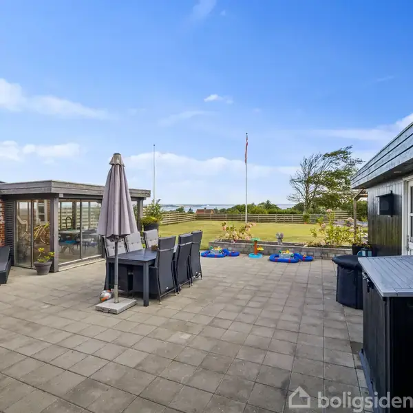 Terrasse med havemøbler og parasol står i midten. I baggrunden ses en flagstang og græsplæne med buske og træer. To bygninger omkranser terrassen, en murstens- og en træbygning.