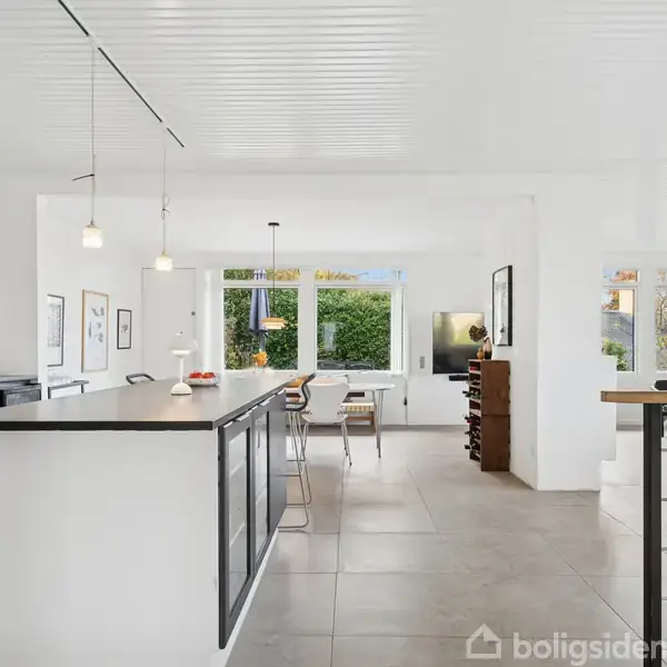 Et moderne køkken med sort-hvide elementer og en stor køkkenø. Ved siden af køkkenet er der en spisestue med lysindfald fra store vinduer og udsigt til en have.