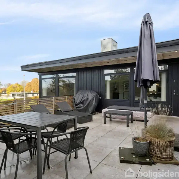 Terrassebord med stole står på flisebelagt terrasse. Ved siden af er solsenge og en grill med overtræk. Parasol og plantekasser bidrager til et hyggeligt, afskærmet haveområde.