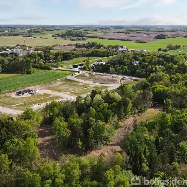 En skov omgiver et byggepladsområde, hvor veje afgrænser tomme parceller. Længere borte ses adskillige landbrugsejendomme og marker under en blå himmel med spredte skyer.