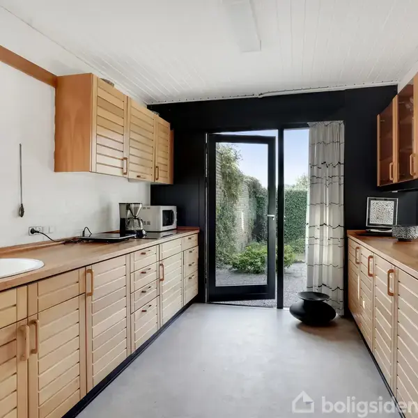 Et køkken med træskabe langs begge sider, en vask til venstre og en mikrobølgeovn. En glasdør fører ud til en have, mens vinduer slipper lys ind.