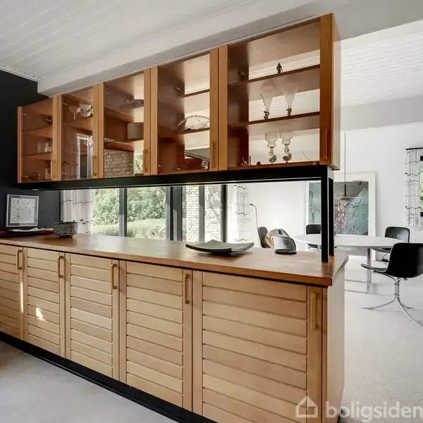 Et træskab står langs en væg, med glasvitrine ovenpå indeholdende dekorative genstande. I baggrunden ses en spisestue med sorte stole og et bord, omgivet af store vinduer med lysindfald.